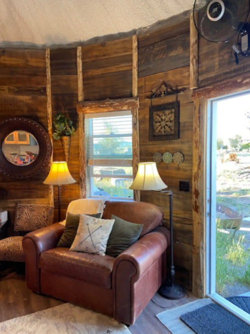 Cozy living space with a brown leather couch, wooden walls, lamps, and a door leading to an outdoor view.